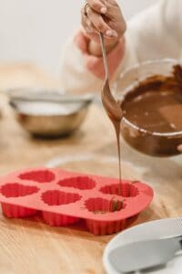 Crop anonymous African American female adding sweet delicious melted chocolate in red heart shaped silicone baking dish
